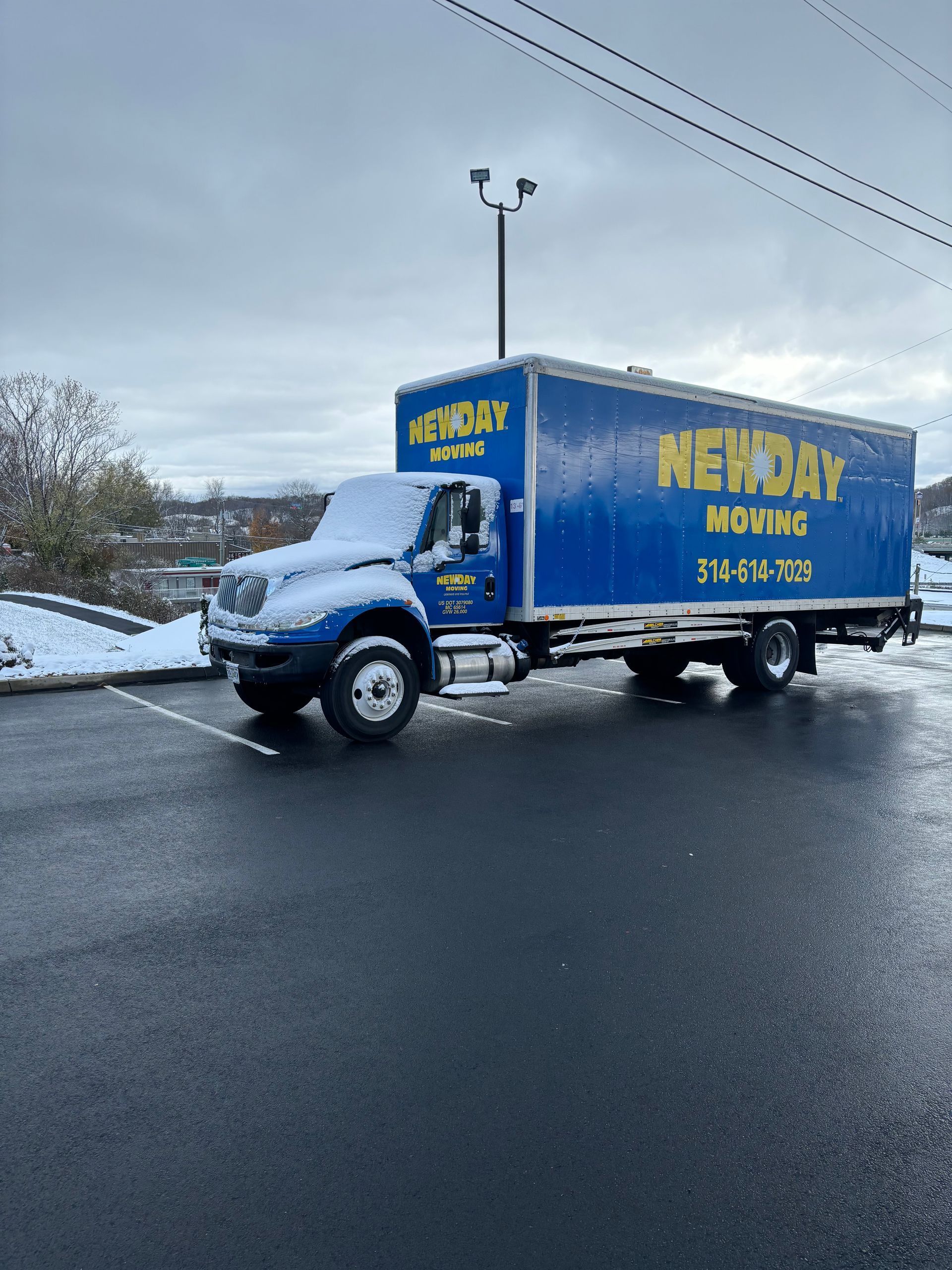 Blue moving truck with yellow lettering driving on a snowy road under a gray sky