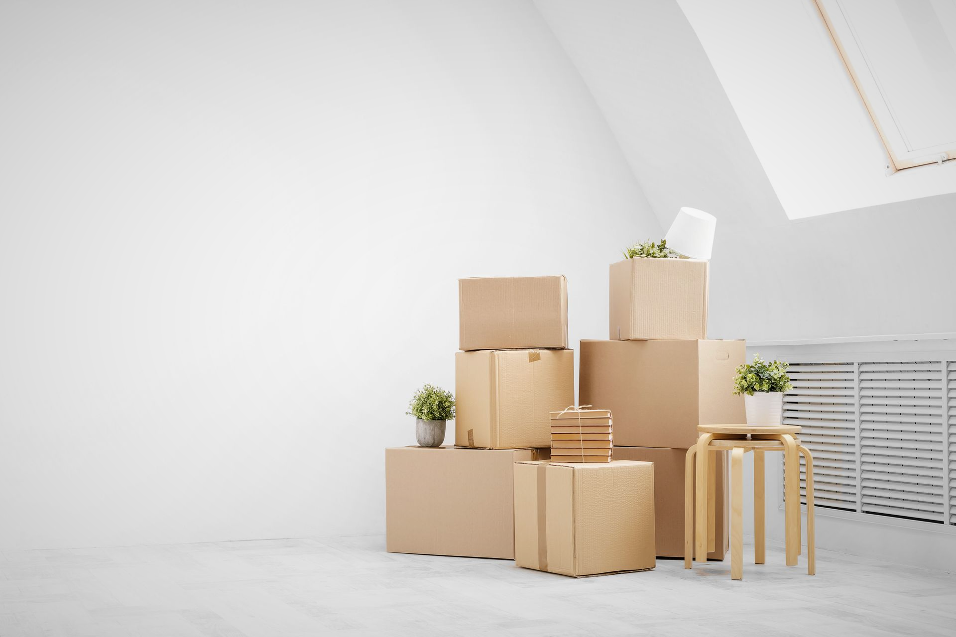 Cardboard moving boxes stacked in a bright, white room with small plants and a wooden stool.