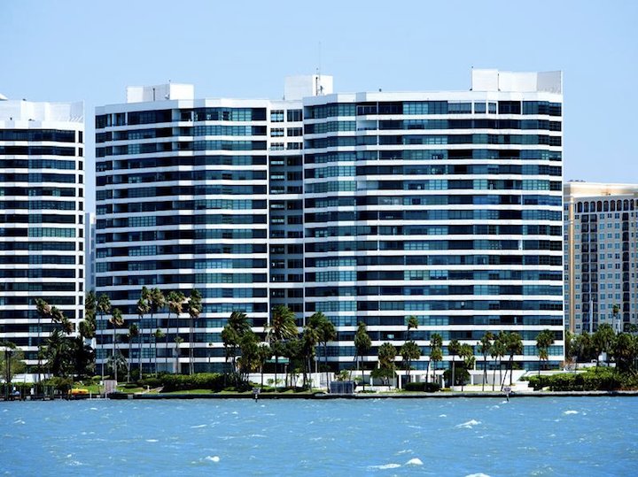 Modern waterfront high-rise condos with blue glass balconies behind palm trees and water