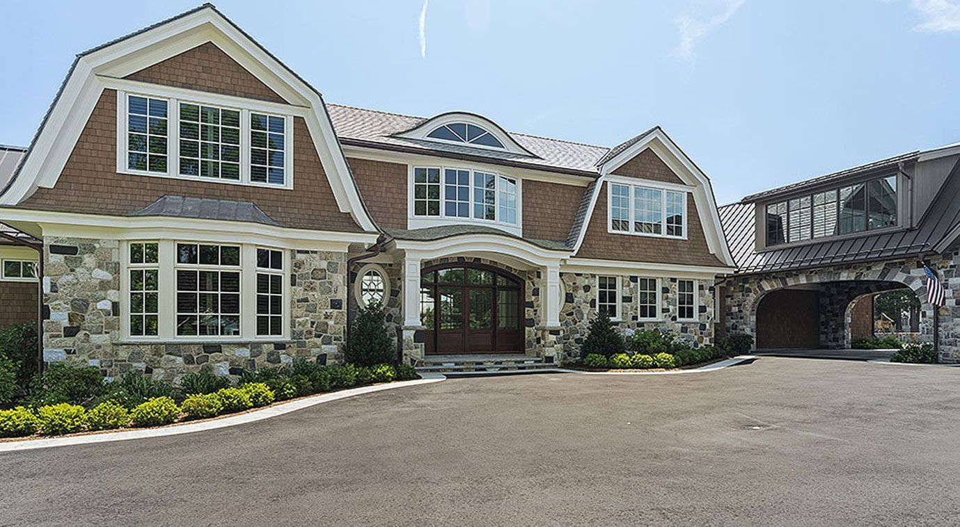 Large stone house with a curved driveway and brown roof, with clear sky visible.