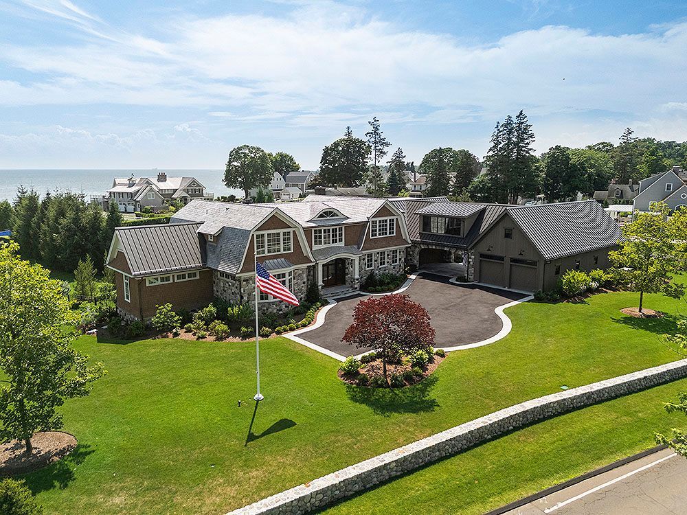 Large brown house with American flag on a green lawn, overlooking the ocean.