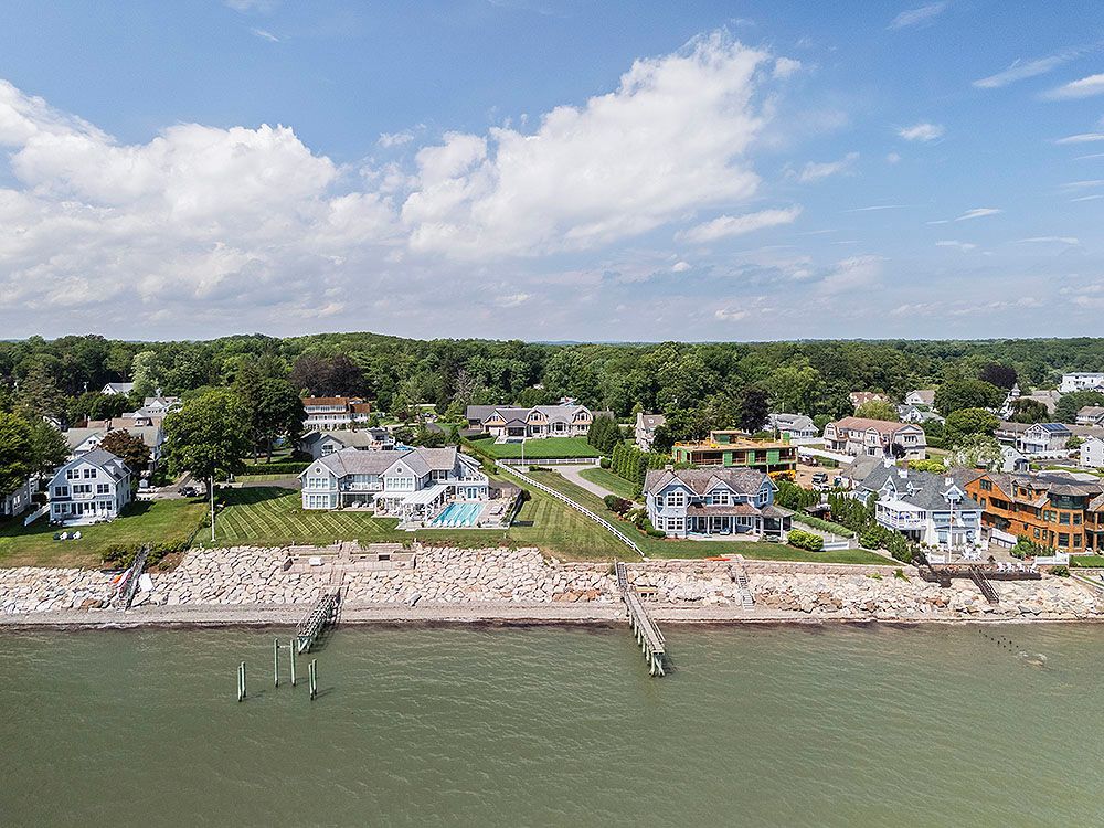 Aerial view of waterfront homes along a shoreline, with trees and blue sky.