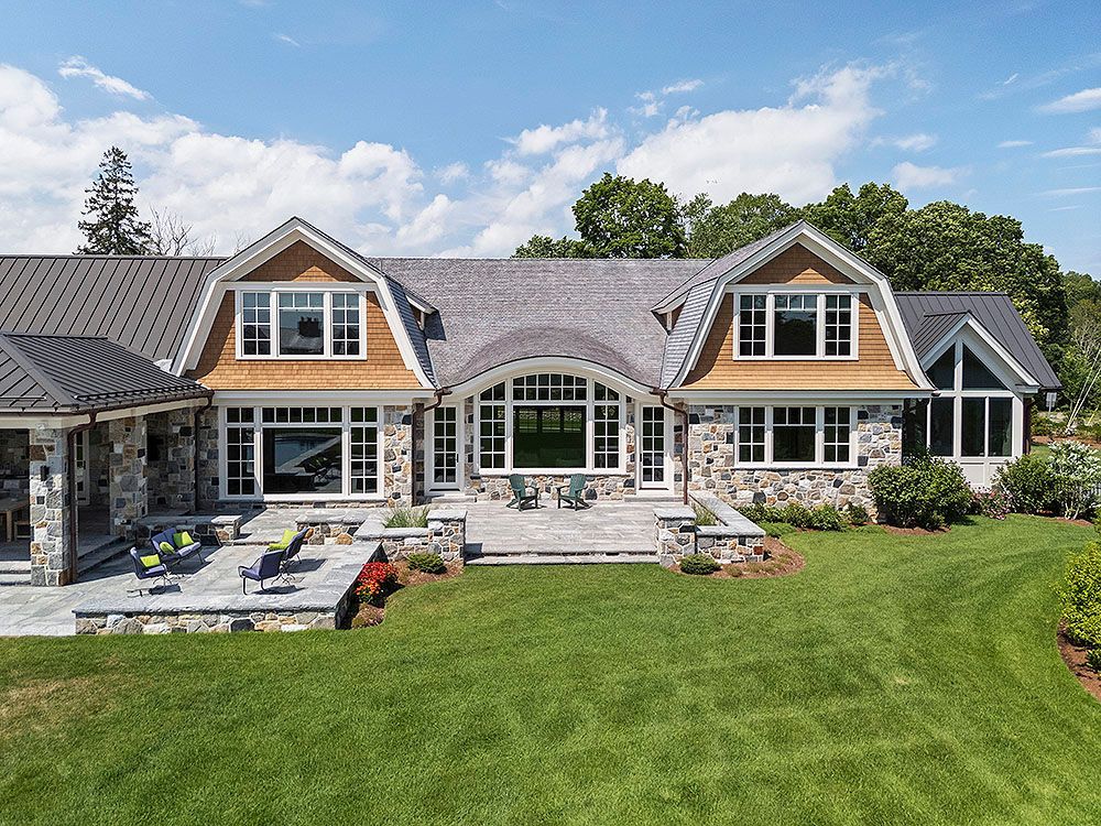 Stone and wood house with dormers, overlooking a green lawn and patio on a sunny day.