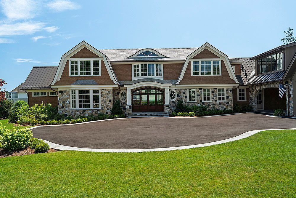 Large stone and brown house with a curved driveway and green lawn under a blue sky.