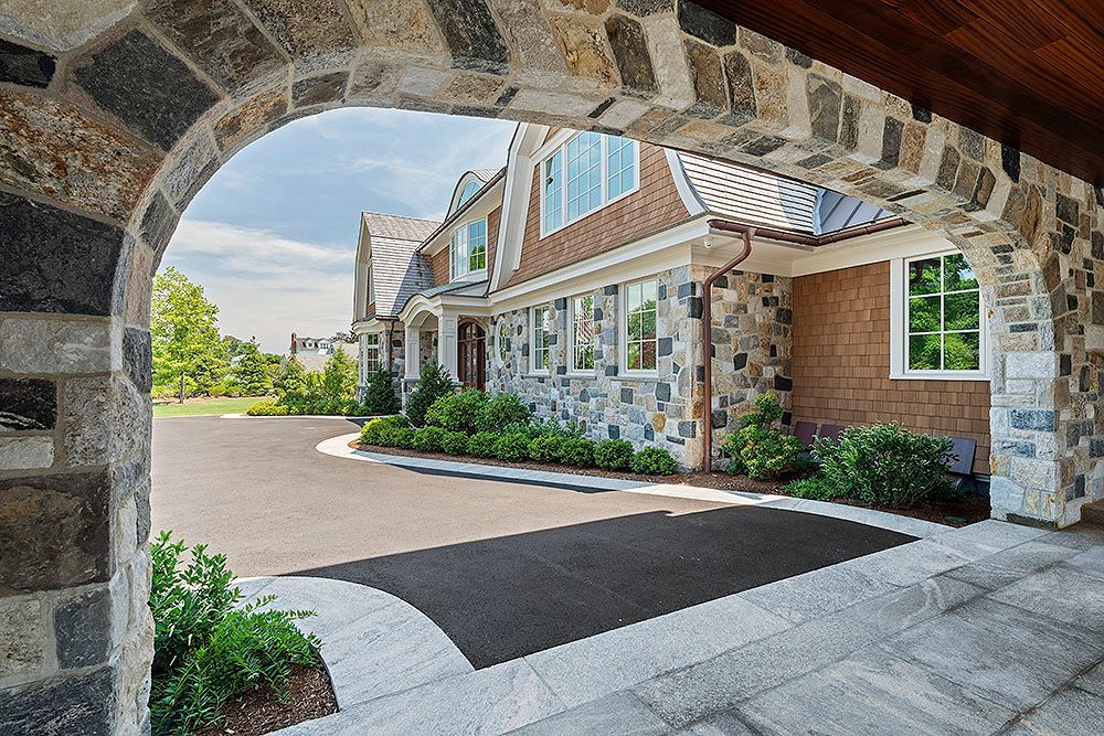 Stone archway frames a luxury house with a gray asphalt driveway, green bushes, and a blue sky.