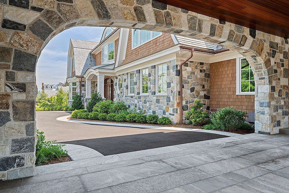 Stone archway frames a house with stone and wood siding, bushes, and a driveway.