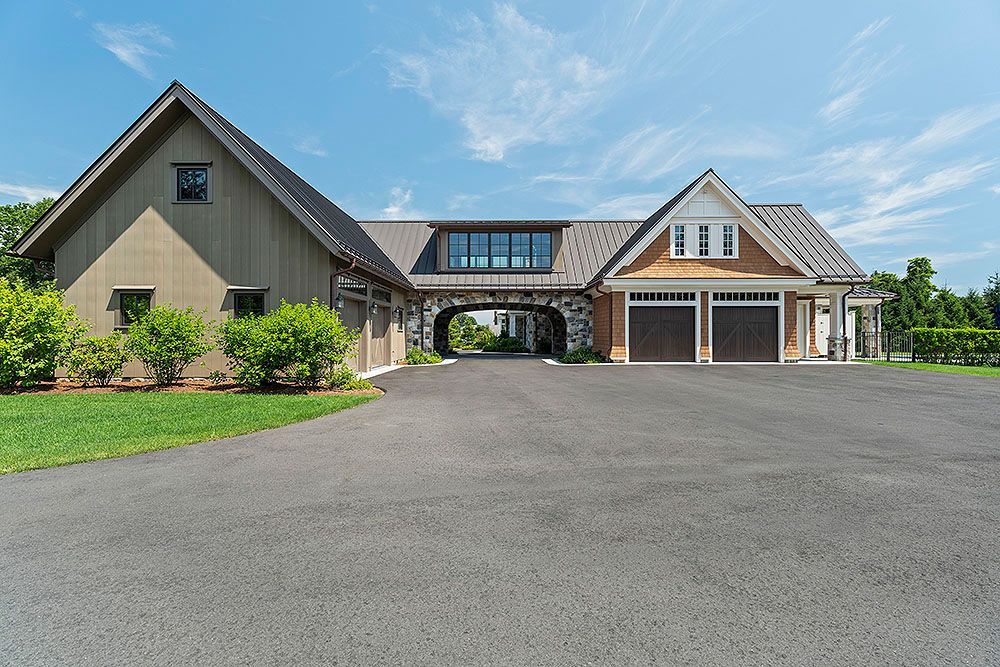 A modern barn-style building with a paved driveway, two garage doors, and a stone archway under a blue sky.