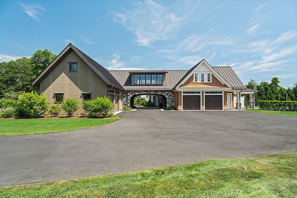 A long driveway leads to a house with brown and tan siding, two garage doors, and a stone archway.