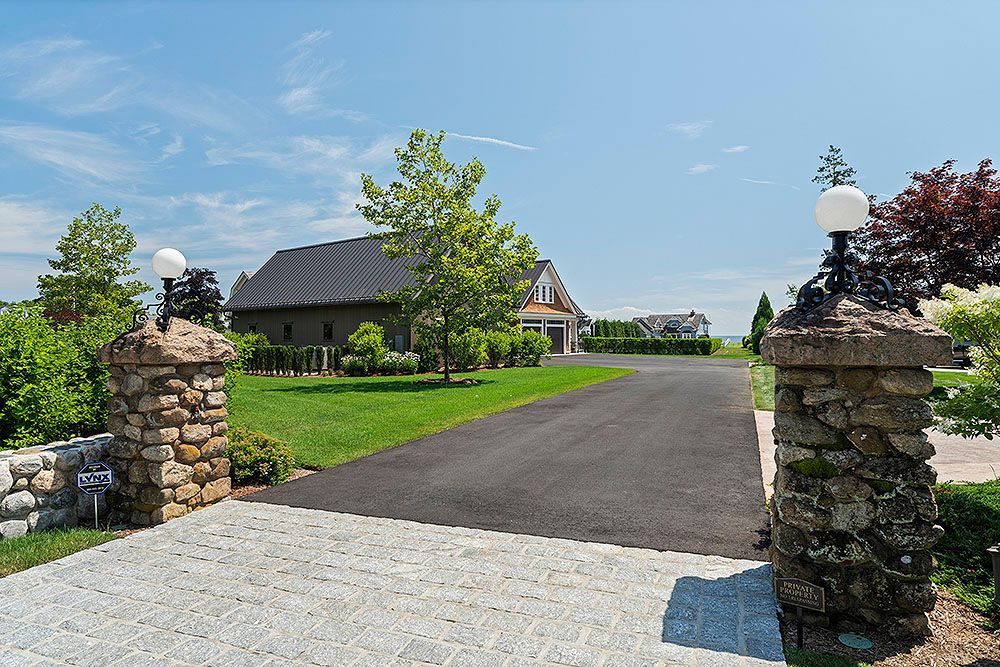 Stone pillars mark the entrance to a driveway leading to a house with a black roof, on a sunny day.