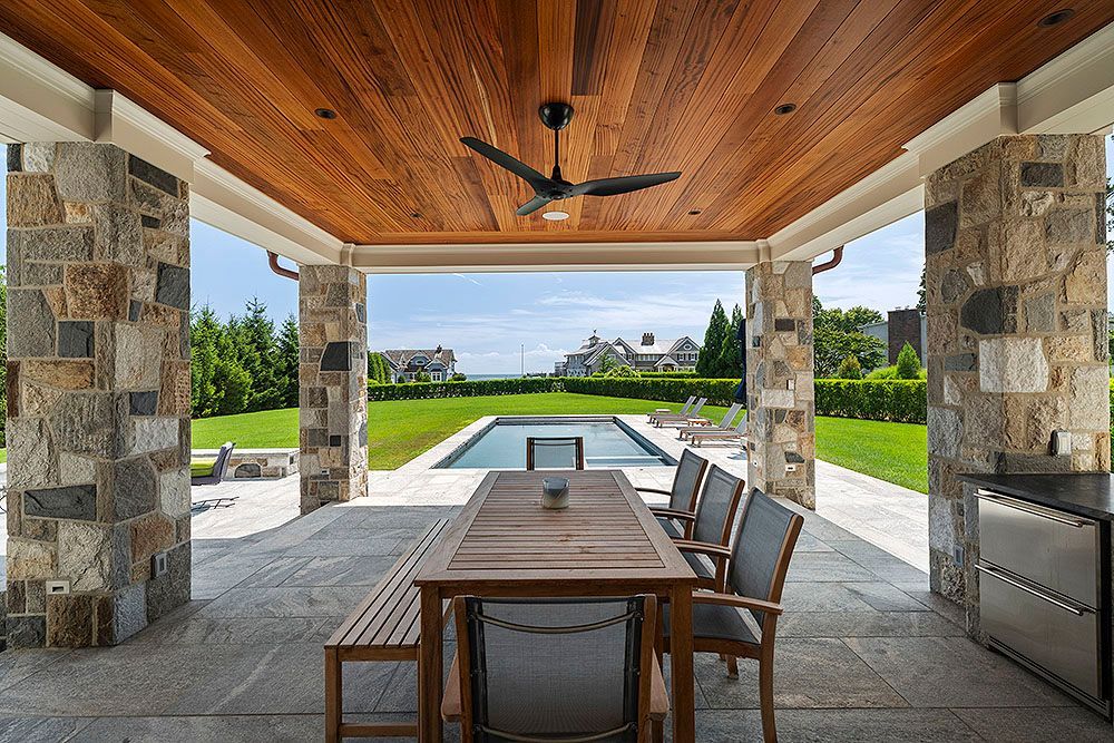 Outdoor dining area with stone columns, wooden ceiling, table, and chairs overlooking a pool and lawn.