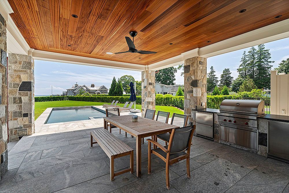 Outdoor kitchen and dining area with stone columns, wood ceiling, and a view of a pool and lawn.