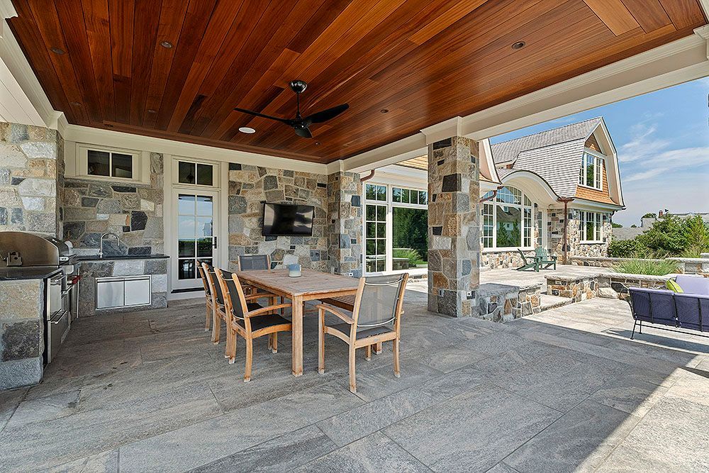 Covered patio with a dining table, grill, stone columns, and a view of a house.