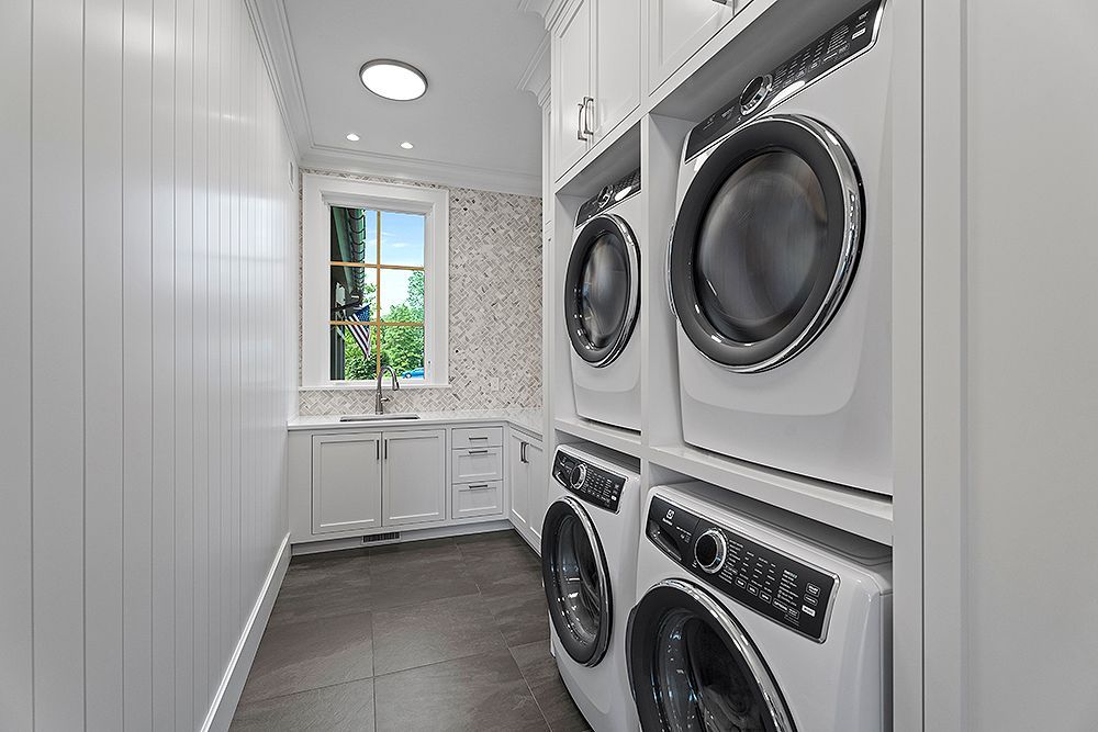 A modern, white laundry room with stacked washer and dryer, sink, and window.