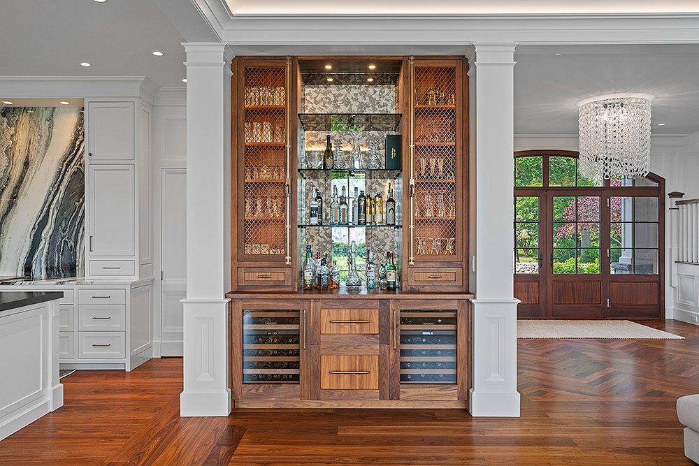 Wooden bar with wine fridges, shelves of liquor, and decorative glass. Between white pillars in a room with hardwood floors.