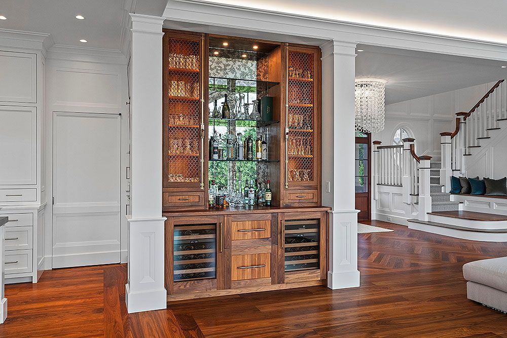 Wooden home bar with liquor display, wine fridges, and open-concept staircase in a bright, modern home.
