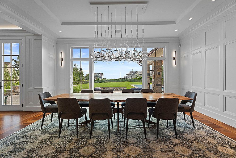 Dining room with large table, chairs, chandelier, large windows, and a view of greenery.