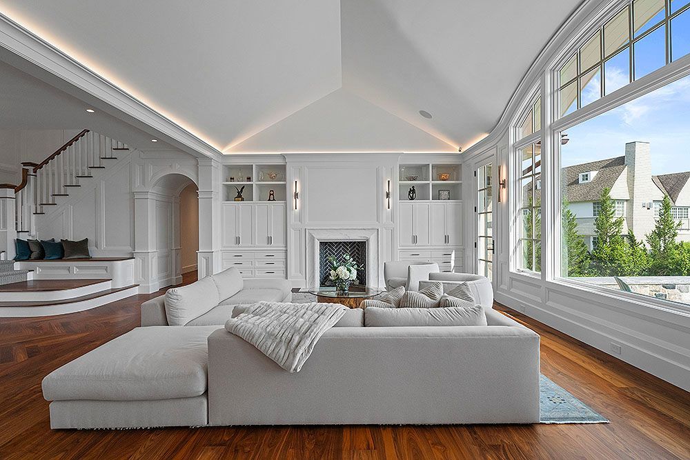 Elegant white living room with a large sectional sofa, fireplace, and arched windows overlooking a neighborhood.