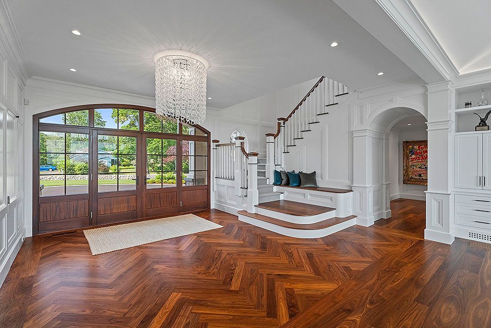 Elegant foyer with herringbone wood floor, large wooden doors, staircase, and chandelier.