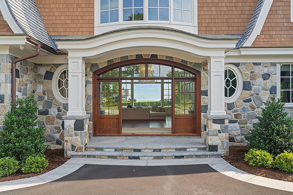 Stone facade home entrance with arched wooden doors, white trim, steps, and landscaping.