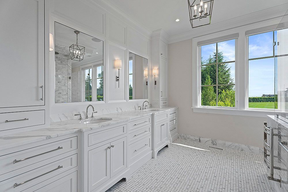 White bathroom with marble countertops, double vanity, large mirrors, and a sunny window with a view.