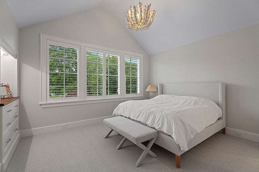 Bedroom with a white bed, windows, and a chandelier. Neutral tones and bright light.