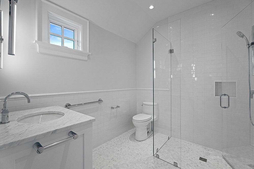 White bathroom with glass shower, toilet, sink, and small window with blue sky.