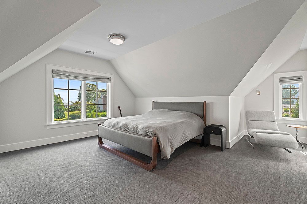 Bedroom with angled ceilings, large windows, bed, and armchair. Grey carpet and white walls.