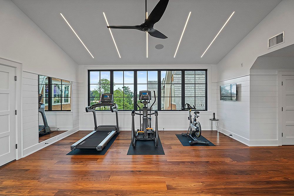 A modern home gym with wood floors, exercise equipment, large windows, and white walls.