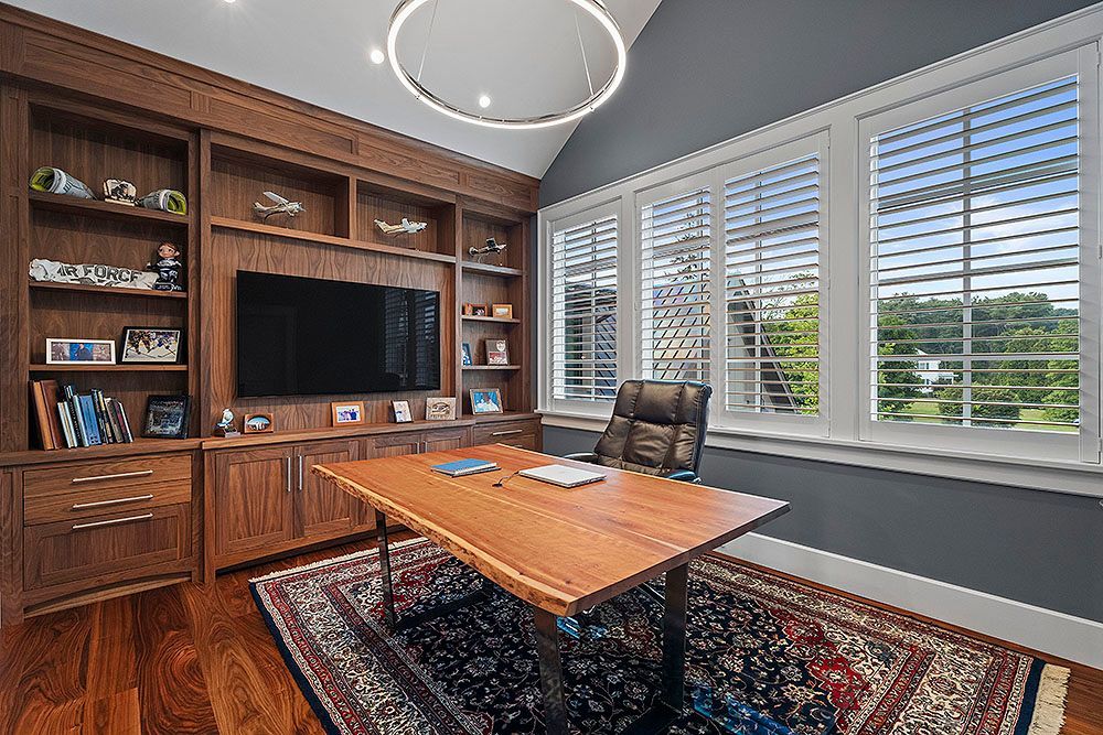 Home office with wooden bookshelves, desk, and window with shutters, rug on hardwood floor.