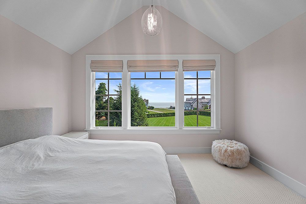 Bedroom with white bed, three-pane window overlooking green yard and blue sky, white walls.