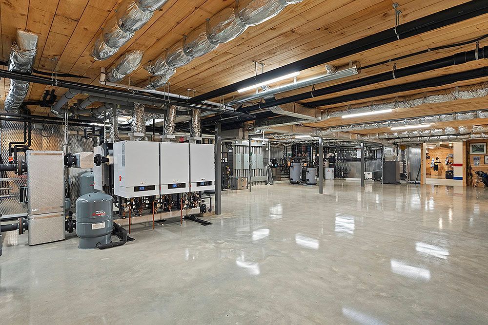 Interior view of a mechanical room with boilers, pipes, and ductwork. Concrete floor, exposed wooden ceiling.