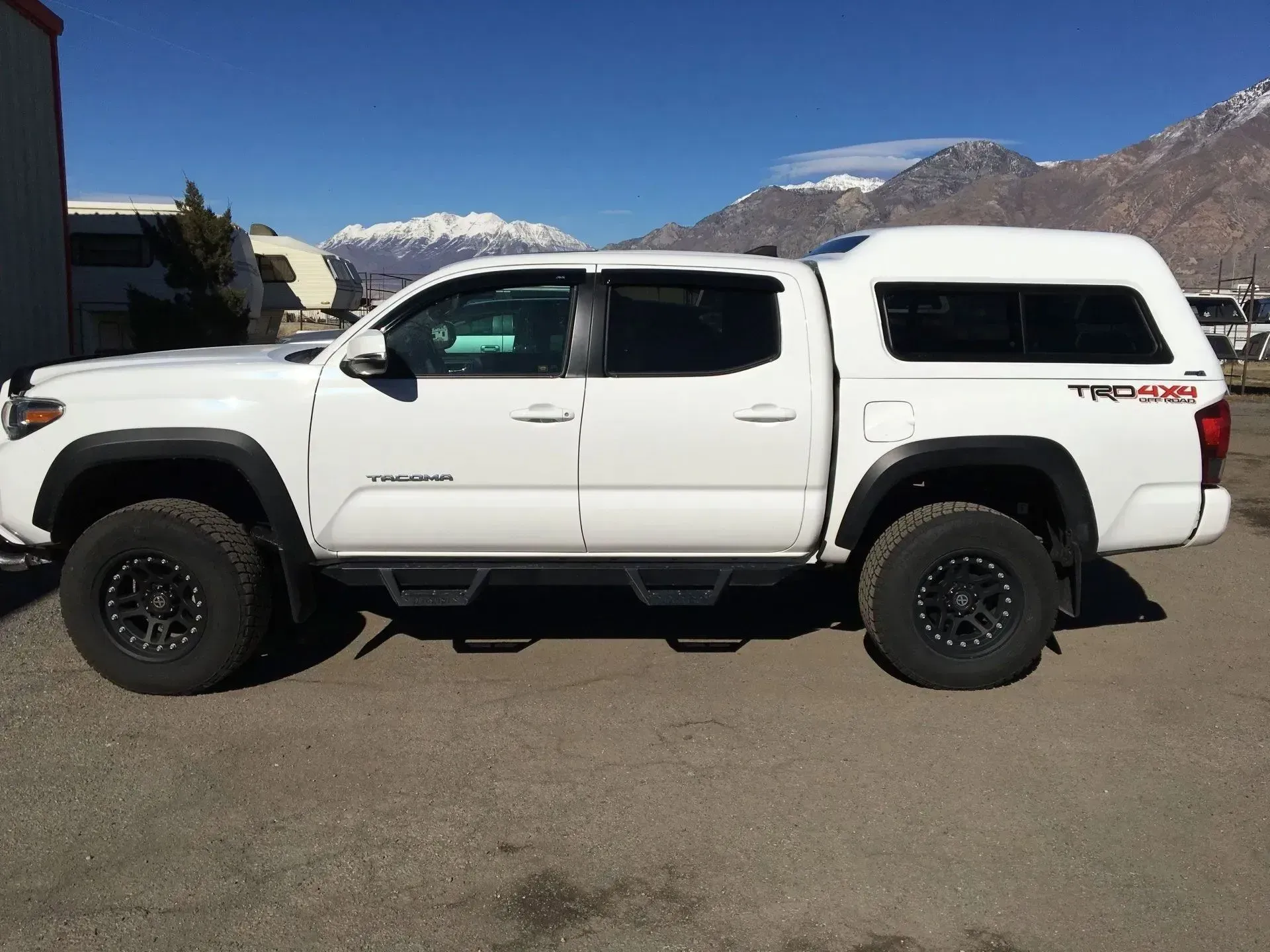 White Toyota Tacoma truck with black wheels and a camper shell, parked on asphalt. Mountains in the background.