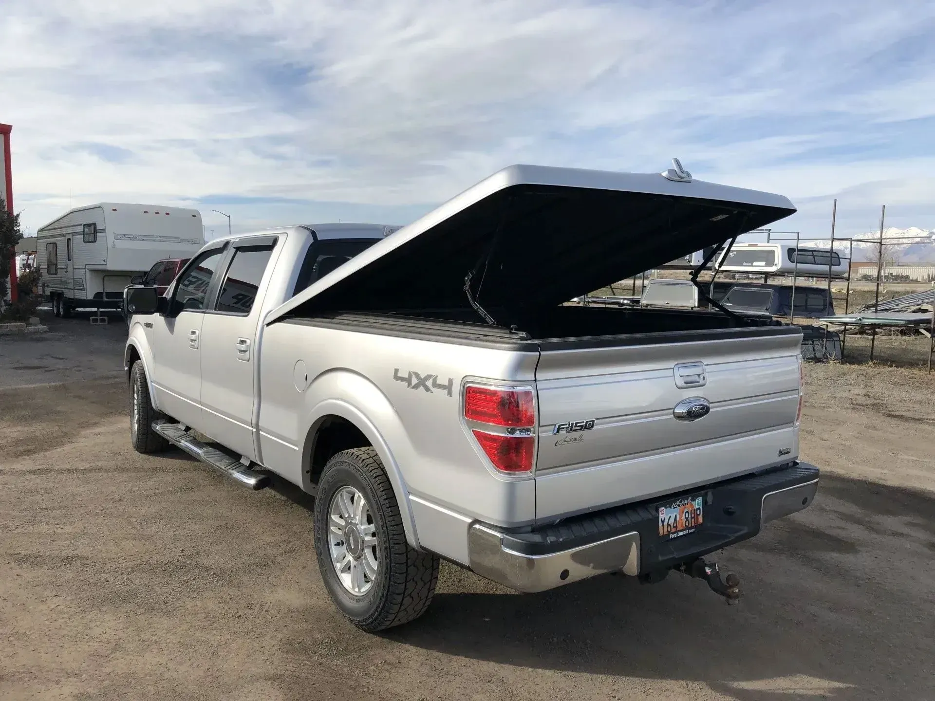 Silver Ford F-150 pickup truck with black bed cover open, parked on dirt lot under a cloudy sky.