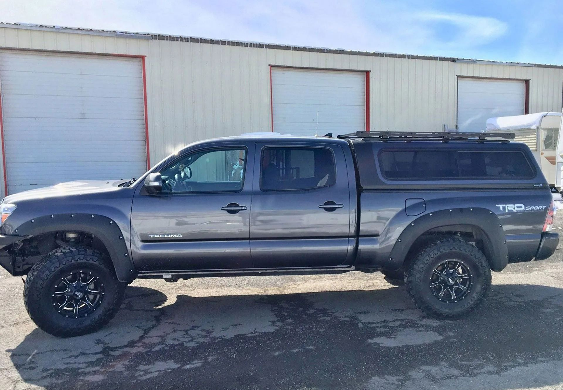 Gray Toyota Tacoma truck with a black cap, black wheels, and roof rack parked in front of a building.