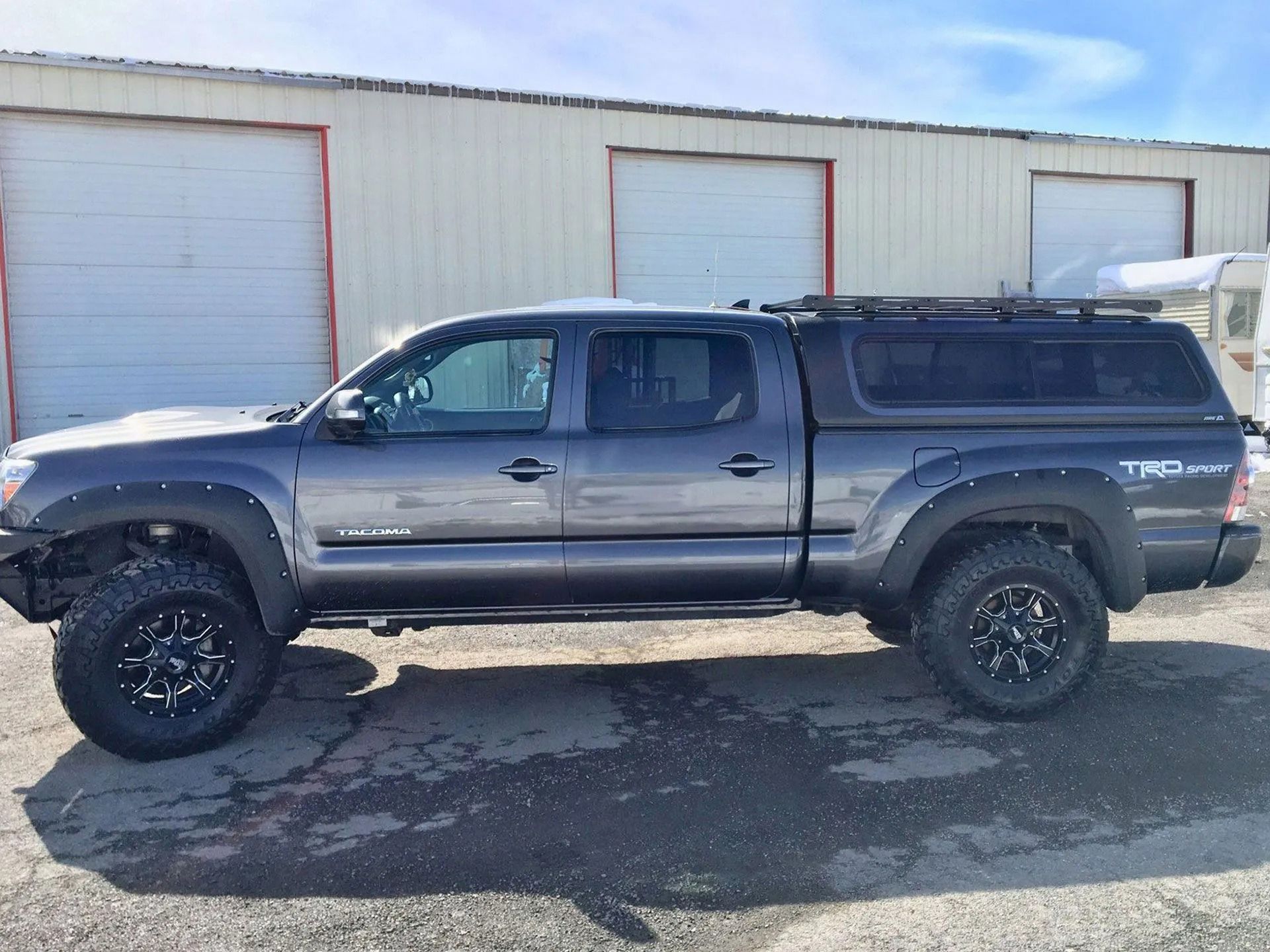 Gray Toyota Tacoma truck with black wheels and a camper shell, parked in front of a building with garage doors.