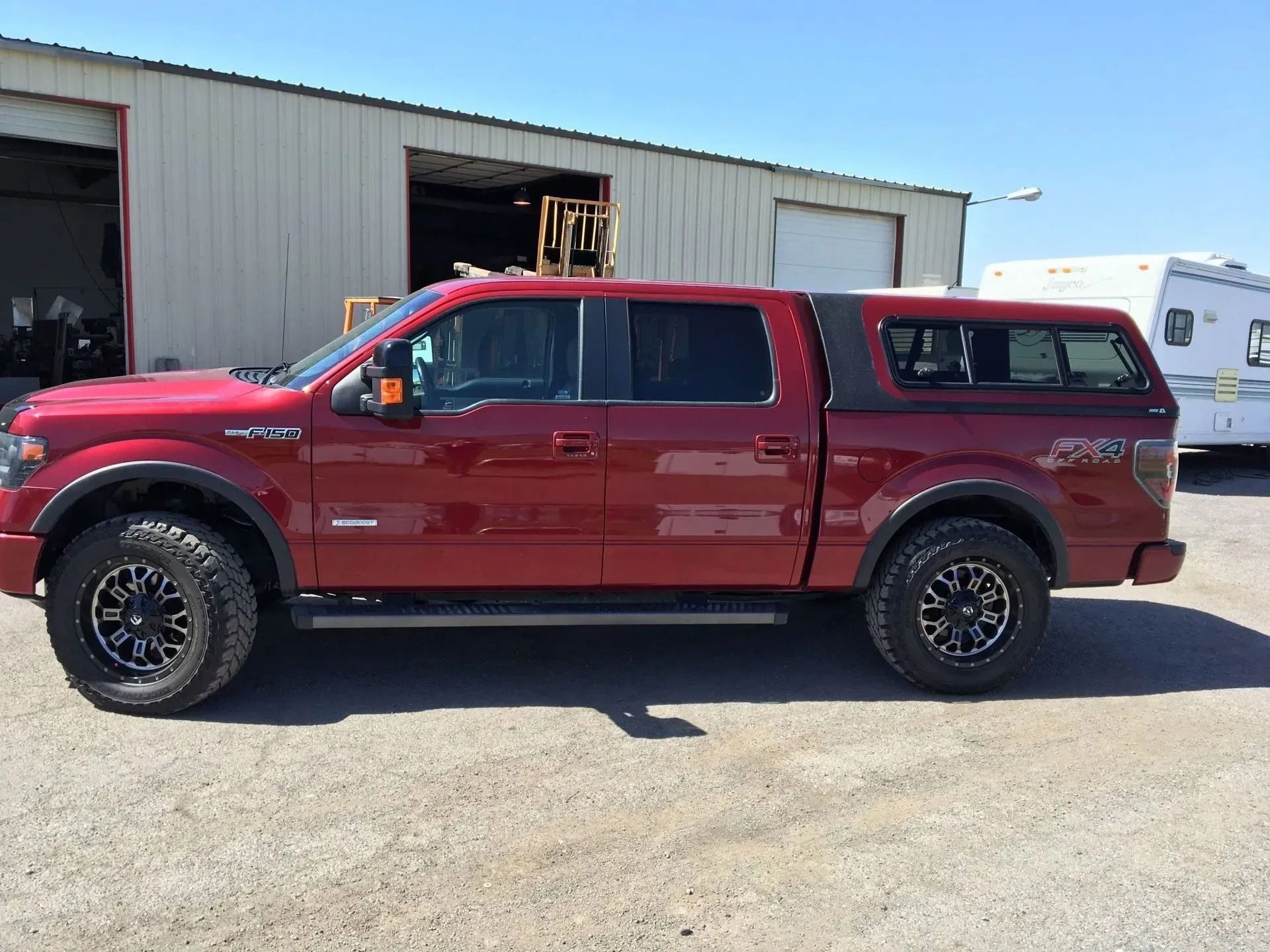 Red Ford F-150 pickup truck with a black cap, black wheels, parked in front of a metal building.
