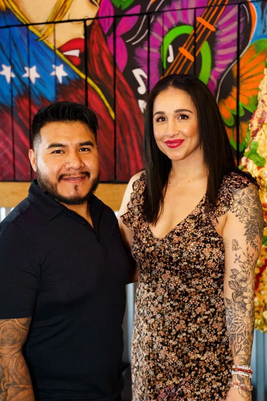 Man and woman smiling, posing in front of a colorful mural. They appear to be in a restaurant or bar.