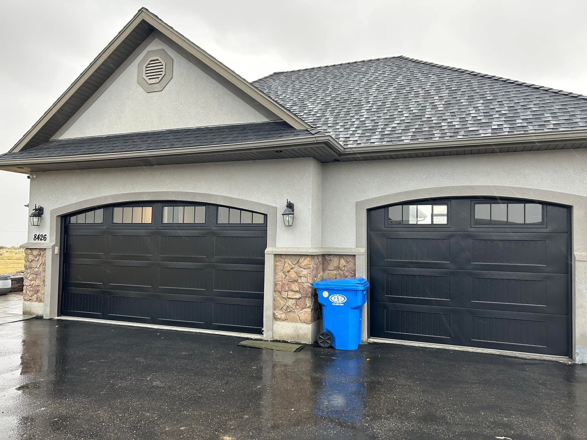 Two-car garage with black doors, light tan stucco, and blue recycling bin on a rainy day.