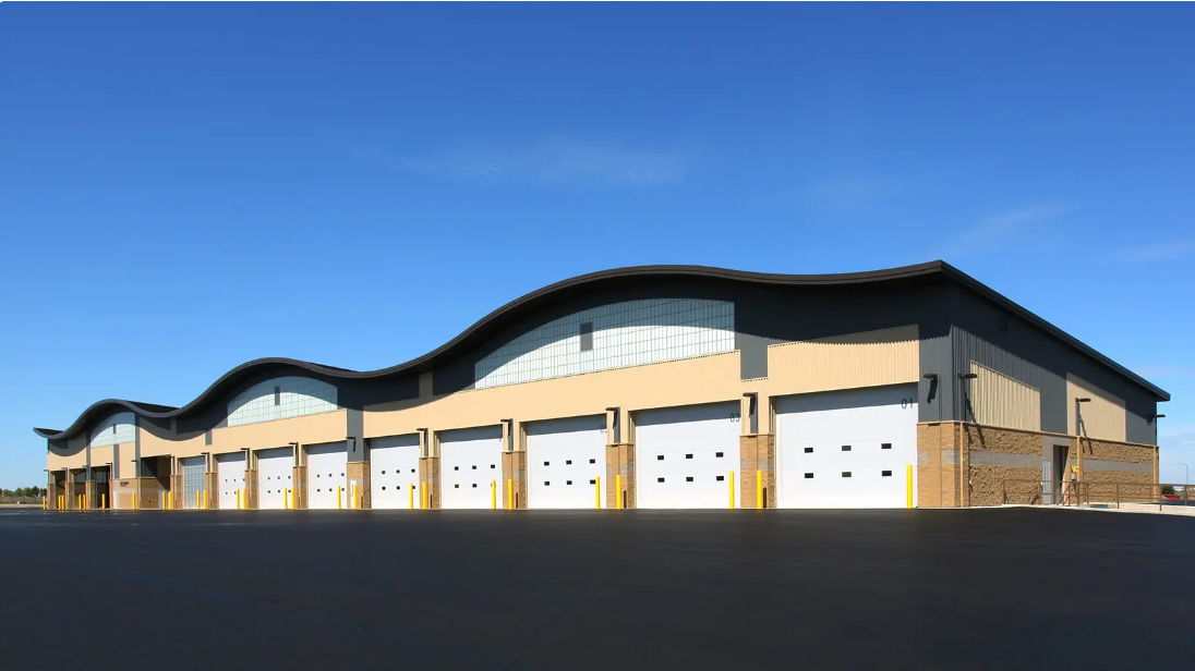 Tan warehouse with wavy roof, several white garage doors, set against a clear blue sky.