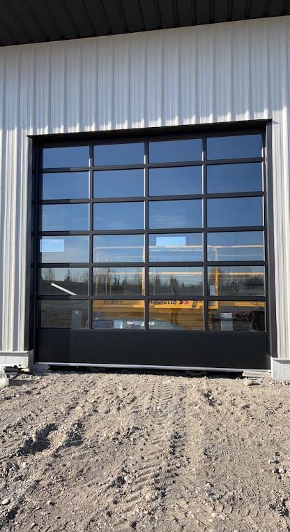 Glass garage door with black frame on a white metal building; gravel in foreground.