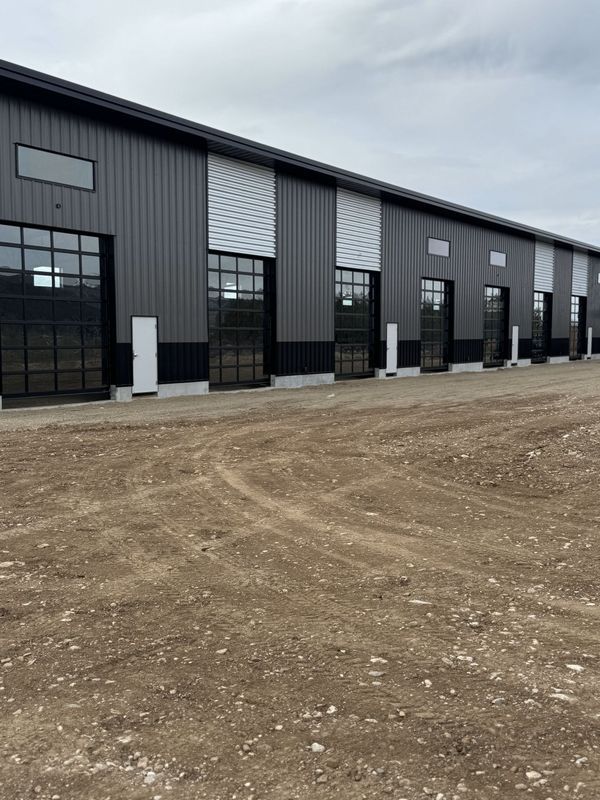 Long, dark gray industrial building with glass garage doors on a gravel lot under a cloudy sky.