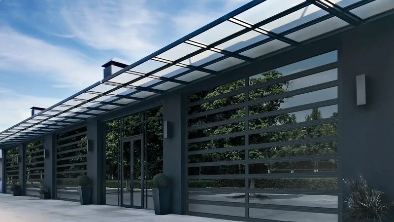 Modern gray gate with glass panels and a glass canopy, set against a sky with trees in the background.
