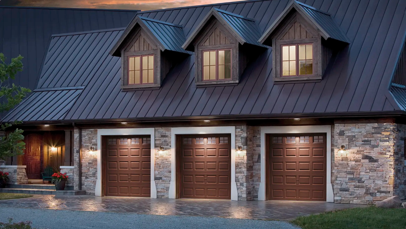 Three brown garage doors on a stone house with dormer windows and a metal roof.