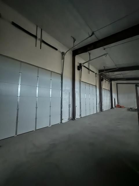 Empty warehouse interior with multiple closed white garage doors, concrete floor, and exposed beams.
