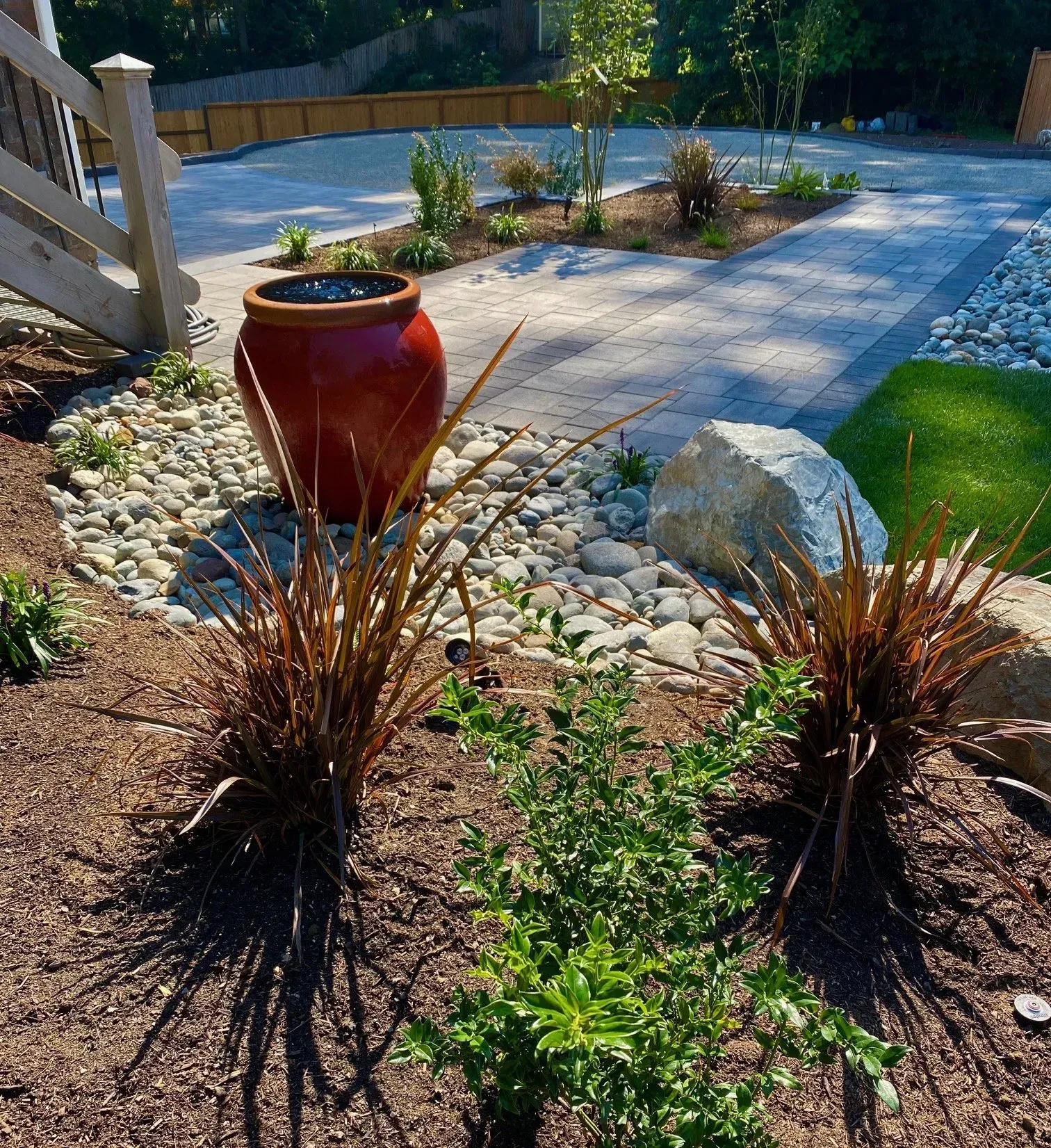 Landscaped yard with a red pot fountain surrounded by rocks and plants, paved pathway in the background.