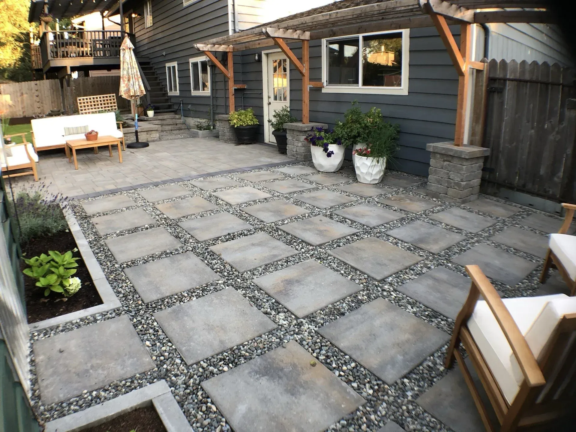 Patio with square concrete pavers, gravel, seating, and a pergola with wooden posts against a gray house.