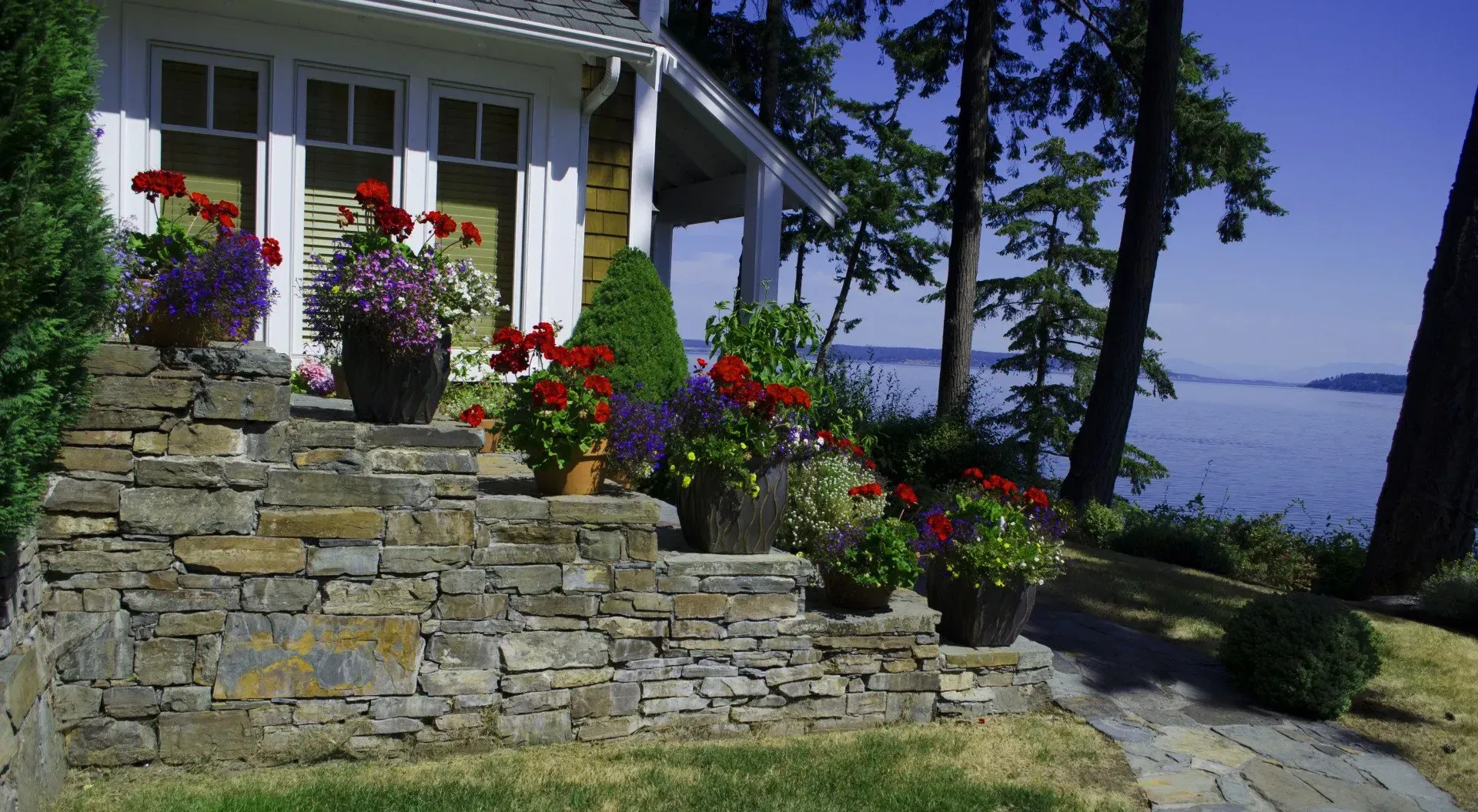 Stone steps lead to a house with potted flowers. Trees and water in the background on a sunny day.