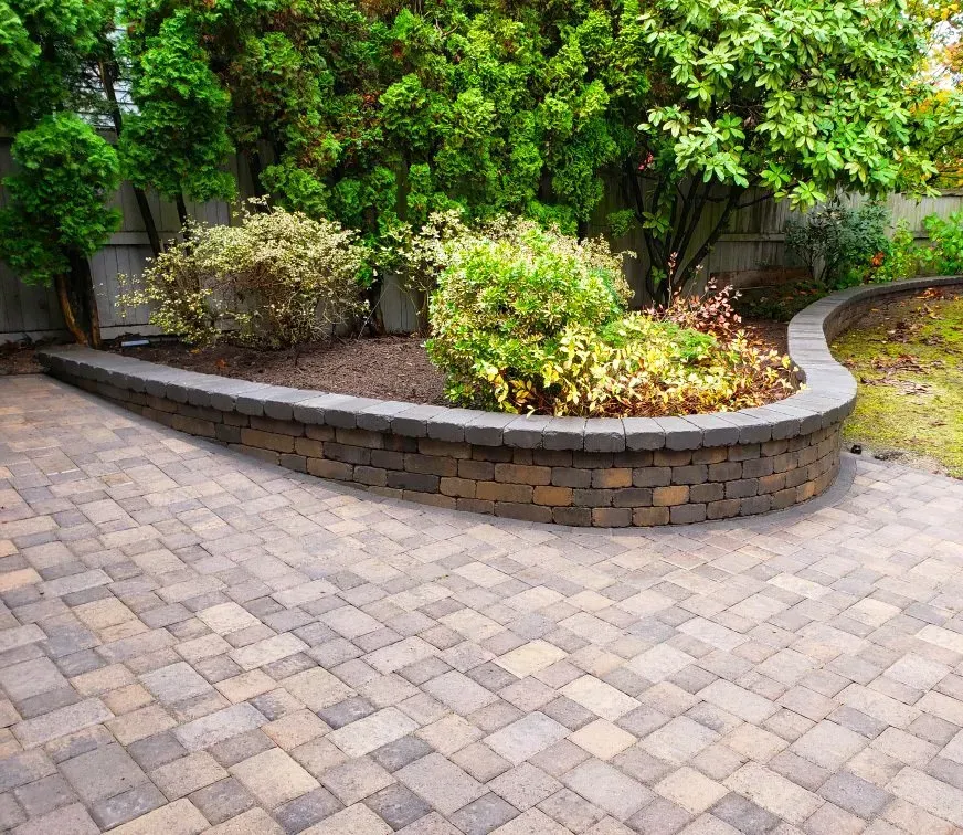 Brick patio with a curved retaining wall bordering a garden bed with green shrubs.