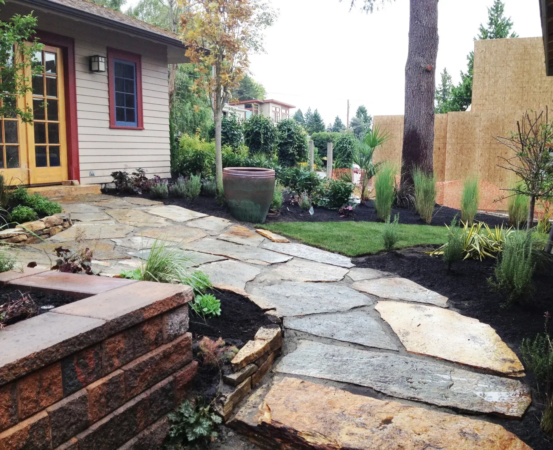 Flagstone path in a landscaped yard, leading to a building with tan siding, greenery, and a large tree.