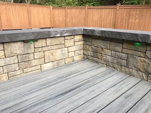 Stone wall with gray composite deck and wooden fence in background; green lights.